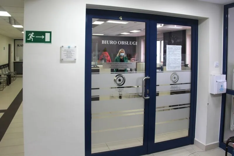 Dwuskrzydłowe drzwi z napisem "BIURO OBSŁUGI" oraz osoba nosząca maseczkę stojąca za szybą.