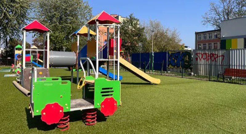 Plac zabaw z kolorowym sprzętem zabawowym, w tym zieloną huśtawką na sprężynach i zjeżdżalnią.