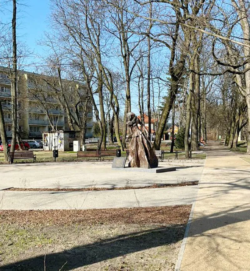 Rzeźba kobiety w sukni na postumencie w parku z ławkami i drzewami bez liści.