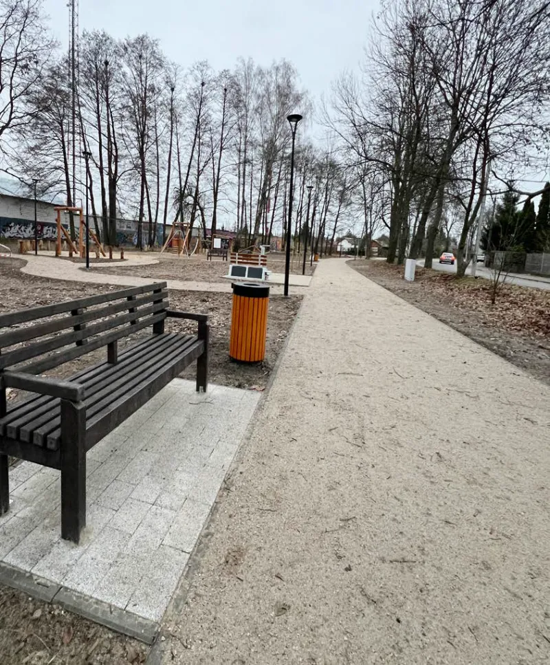 Drewniana ławka i pomarańczowy kosz na śmieci stoją obok szerokiej, utwardzonej ścieżki w parku z gołymi drzewami i placem zabaw w tle.