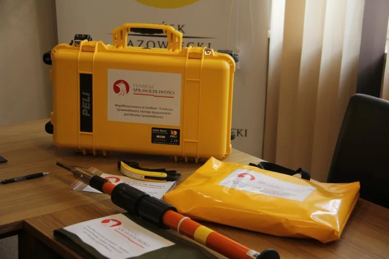 Żółta walizka Peli, pomarańczowo-czarna laska oraz żółta koperta z nalepkami funduszu Sprawiedliwości na drewnianym stole.