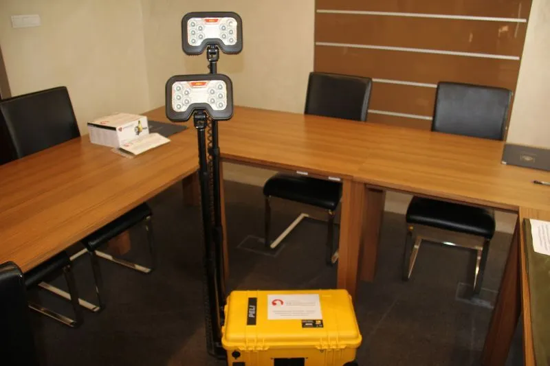 Żółta skrzynka polowa z dwoma zamontowanymi panelami świetlnymi na statywie ustawiona przy drewnianym stole z czarnymi krzesłami.