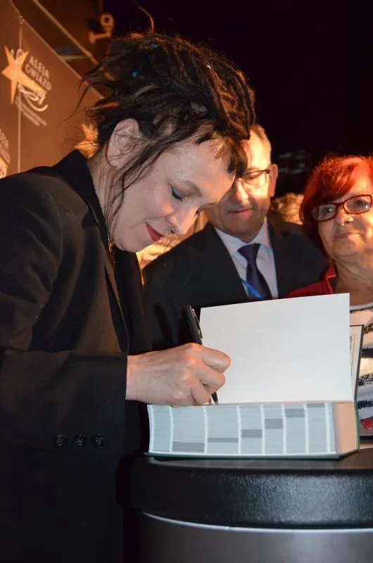 Kobieta z dredami podpisuje książkę na spotkaniu, obok niej stoją dwie osoby w tle.