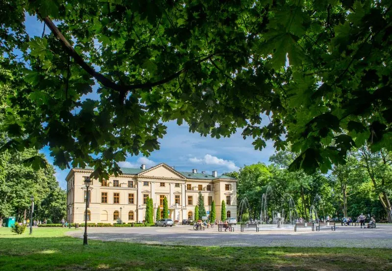 Klasyczny budynek z fontanną i ławeczkami na placu otoczonym drzewami.