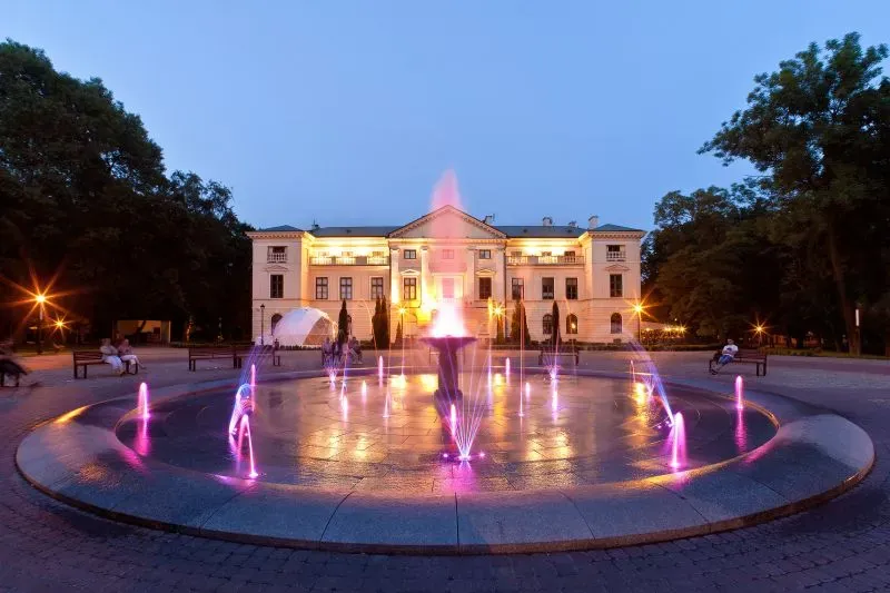 Fontanna z kolorowymi strumieniami wody przed oświetlonym budynkiem klasycystycznym w parku.