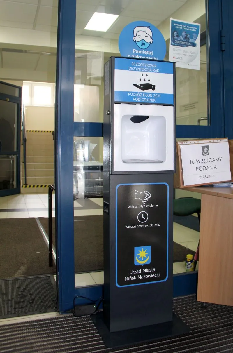 Stojak z bezdotykowym dozownikiem do dezynfekcji rąk z instrukcjami i logo Urzędu Miasta Mińsk Mazowiecki przy wejściu do budynku.