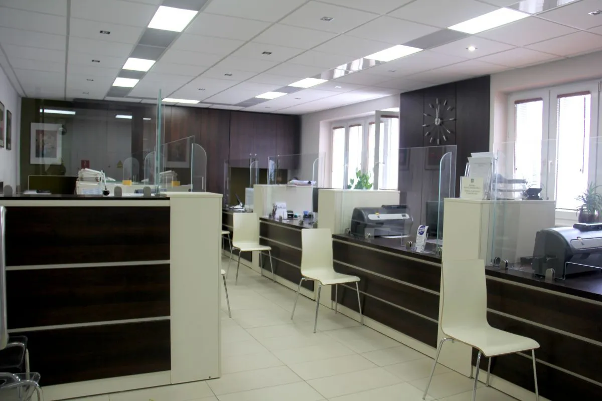 Recepcja z białymi krzesłami, przegrodami ochronnymi z przezroczystego szkła oraz ciemnymi meblami i sprzętem biurowym.