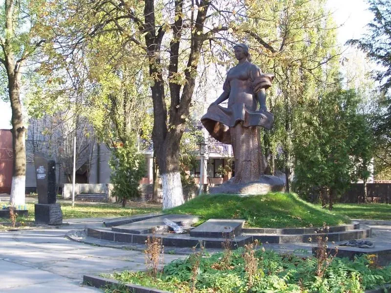 Pomnik kobiety w sukni stojącej na podwyższeniu w parku z drzewami i zielenią.