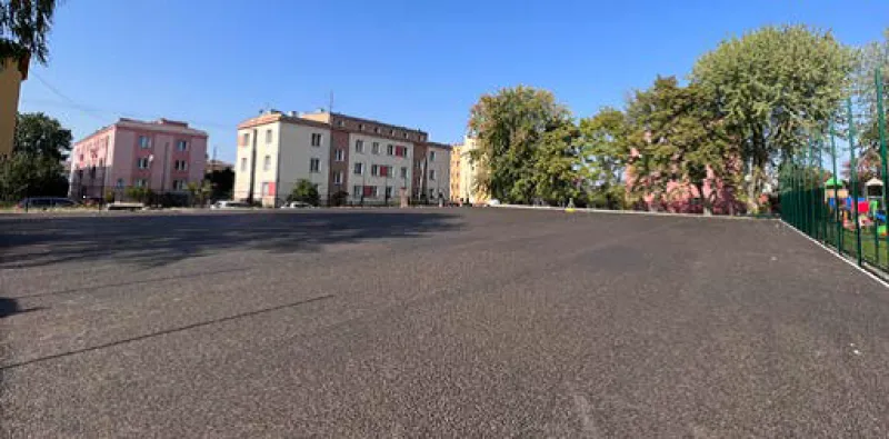 Pusty asfaltowy plac z niskimi budynkami mieszkalnymi i drzewami w tle.