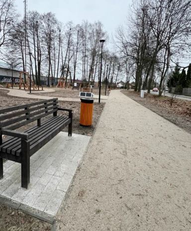 Drewniana ławka i pomarańczowy kosz na śmieci stoją obok szerokiej, utwardzonej ścieżki w parku z gołymi drzewami i placem zabaw w tle.