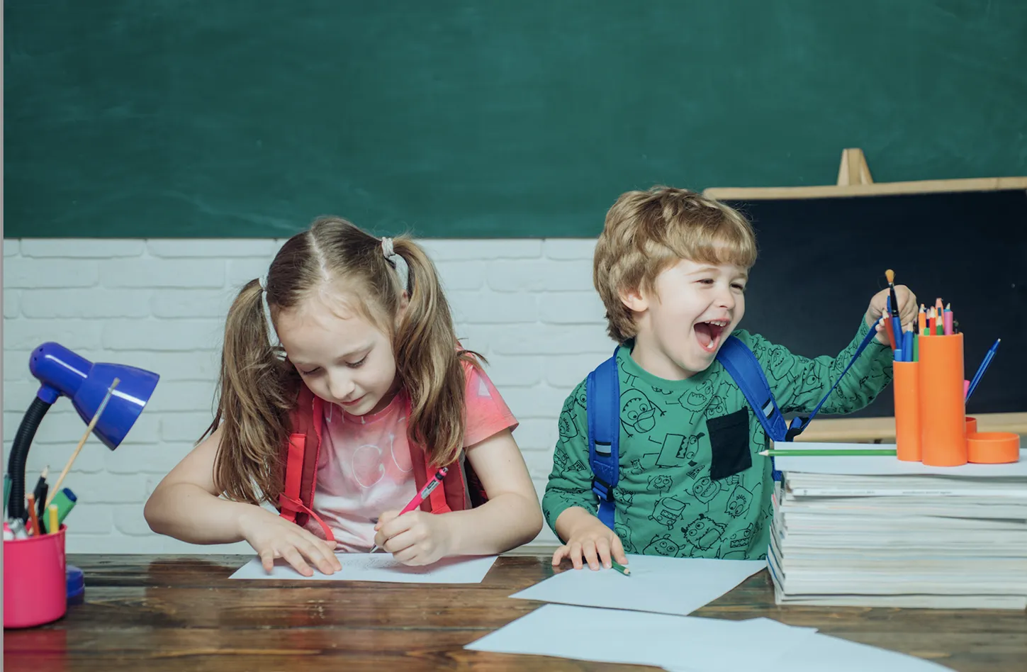 Dwoje dzieci siedzi przy stole z papierem i przyborami do rysowania, jedno pisze, a drugie się śmieje i sięga po kredki.