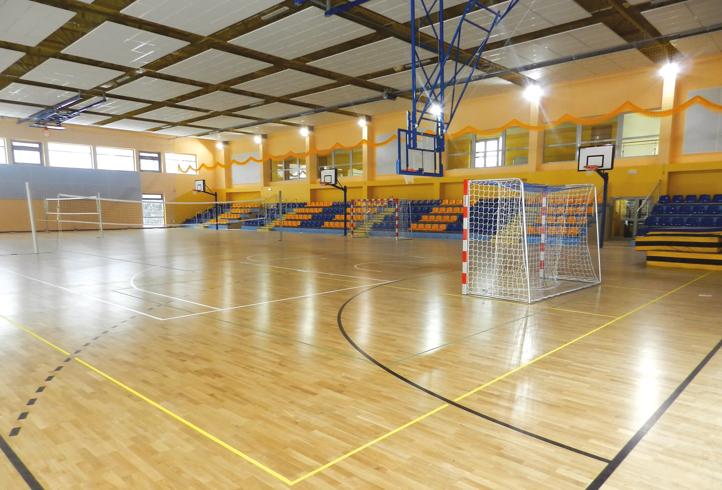 Wnętrze hali sportowej z boiskiem do siatkówki, bramkami do piłki ręcznej oraz trybunami z niebiesko-pomarańczowymi siedzeniami.