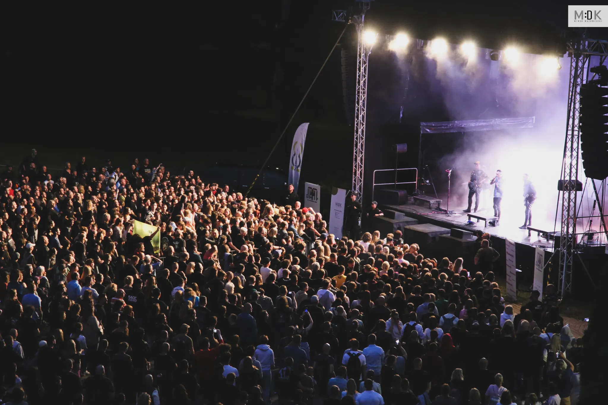 Tłum ludzi ogląda koncert na otwartej scenie z muzykami grającymi przy jasnym oświetleniu.