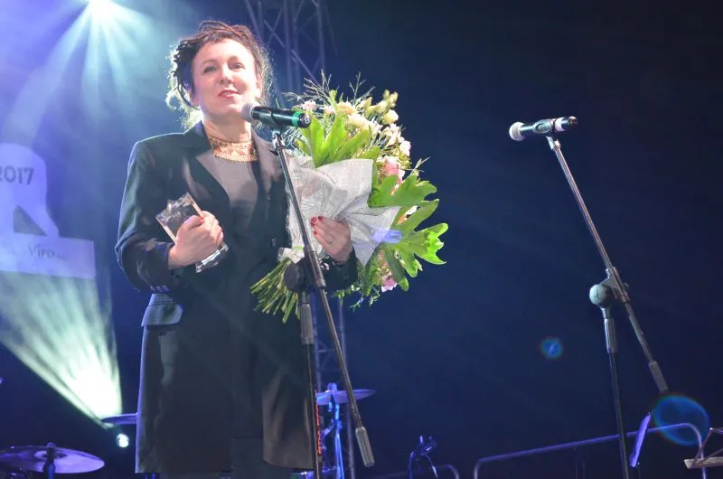 Kobieta w czarnym ubraniu trzyma bukiet kwiatów i statuetkę stojąc przy mikrofonie na scenie.