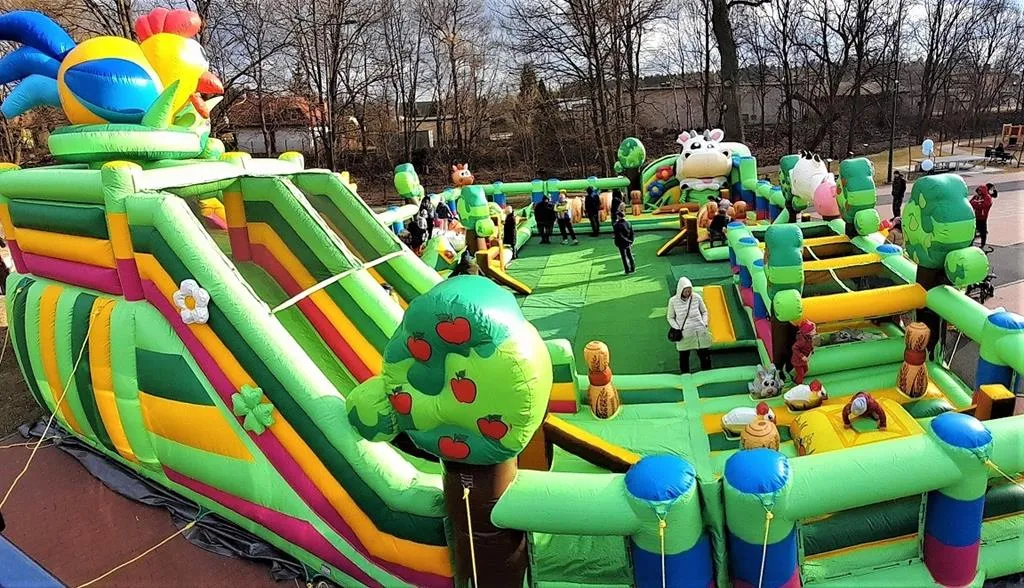 Duży, zielony dmuchany plac zabaw z motywem zwierząt i drzew oraz kilkoma osobami bawiącymi się i chodzącymi po nim.