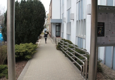 Chodnik obok białego budynku z metalową poręczą i tablicą ogłoszeń, przy którym idzie osoba.
