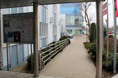 Tablica informacyjna Urzędu Miasta Mińsk Mazowiecki obok budynku z chodnikiem i barierkami.