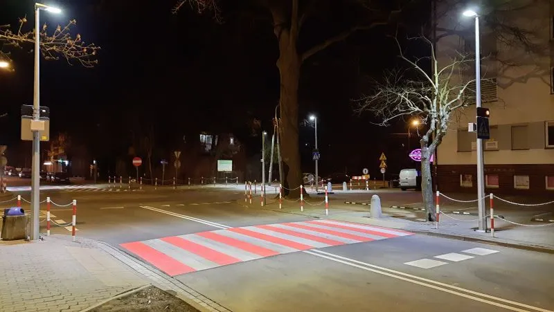 Przejście dla pieszych z czerwono-białymi pasami na oświetlonej ulicy nocą z drzewami i znakami drogowymi.