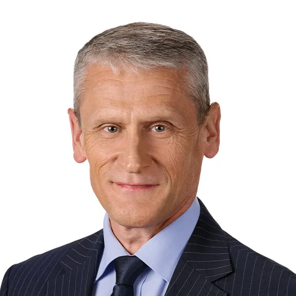 Mężczyzna o krótkich siwych włosach w garniturze w prążki, niebieskiej koszuli i granatowym krawacie.