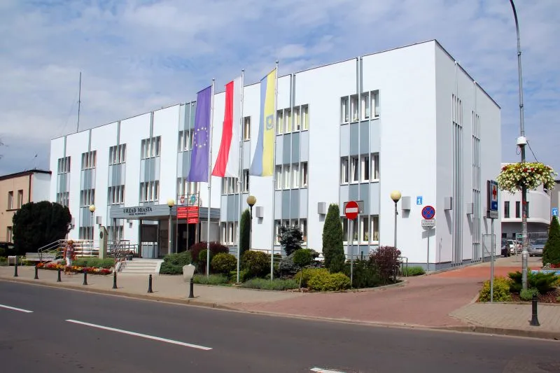 Trzypiętrowy biały budynek z prostokątnymi oknami, czterema flagami na masztach i wejściem oznaczonym napisem „URZĄD MIASTA”.