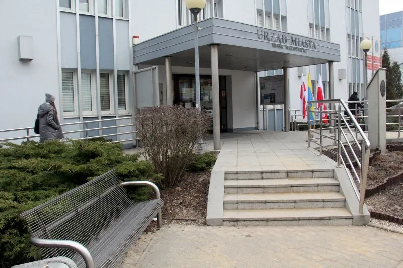 Wejście do budynku Urzędu Miasta z metalową ławką, schodami, podjazdem oraz flagami.