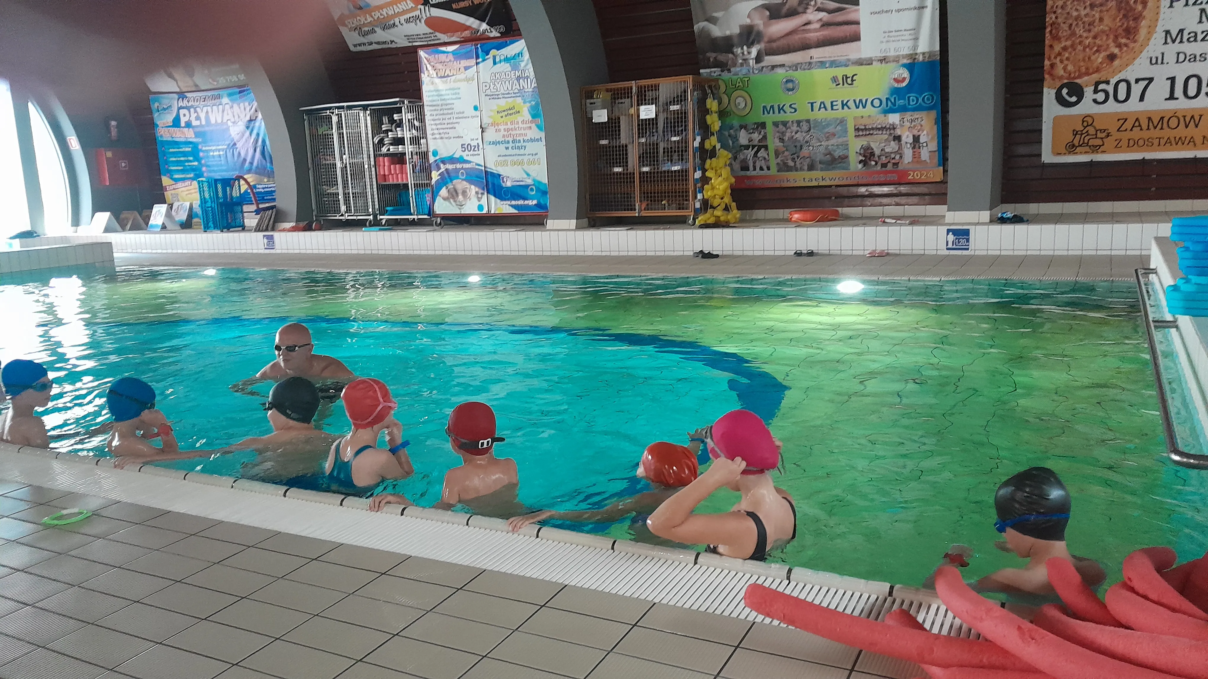 Grupa dzieci w czepkach pływackich stoi w basenie przy krawędzi, mężczyzna w okularach pływackich znajduje się w wodzie naprzeciwko nich, wnętrze z tablicami ogłoszeń na ścianie i sprzętem pływackim.