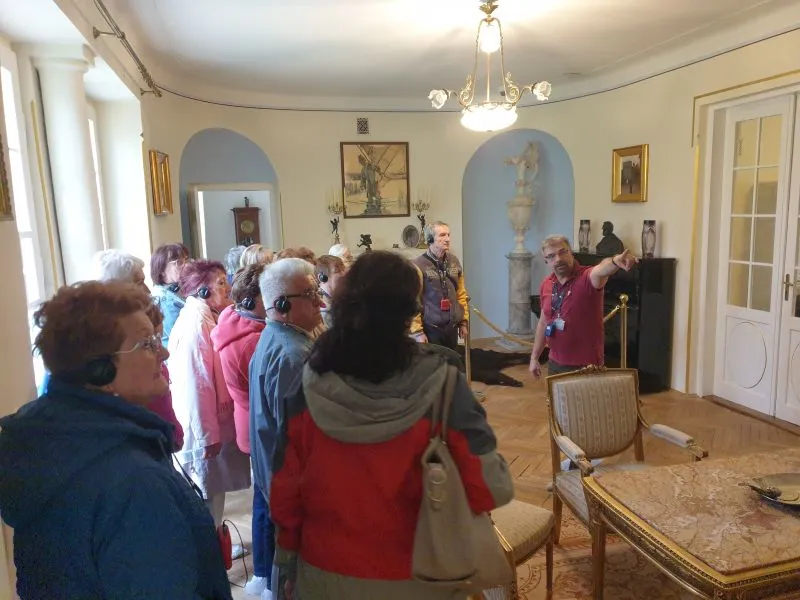 Grupa osób słuchająca przewodnika w historycznym wnętrzu z rzeźbą, obrazami i meblami.