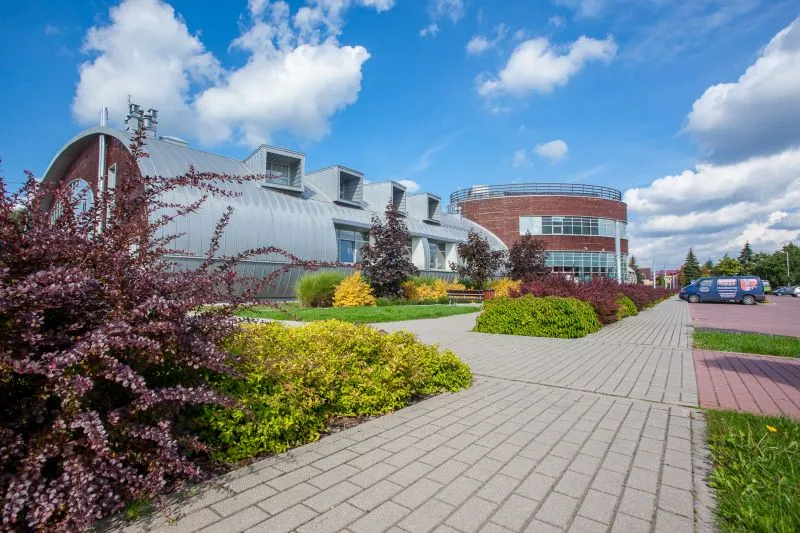 Szary budynek z oknami dachowymi obok okrągłego budynku z czerwonej cegły, otoczone krzewami i chodnikiem z szarych płytek.