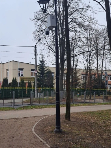 Latarnia uliczna z zamontowaną kamerą monitoringu i drzewami wokół na tle budynku szkoły.