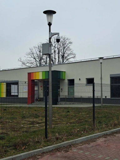 Latarnia z kamerą nad ogrodzeniem na tle budynku z kolorowym wejściem.