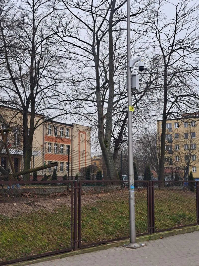 Latarnia z zamontowaną kamerą monitoringu stoi przy ogrodzeniu z siatki, za którym widać drzewa bez liści i budynki mieszkalne.