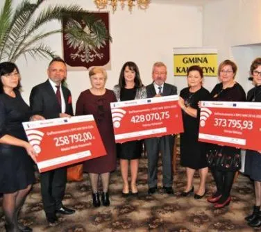 Grupa osób stoi w pomieszczeniu, trzymając trzy duże czeki z widocznymi kwotami i nazwą „Gmina Kamień”.