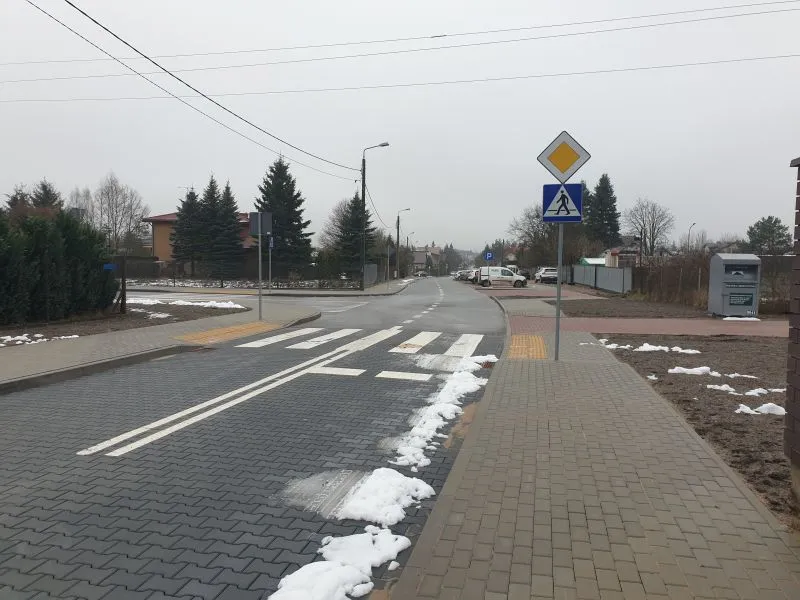 Ulica z pasami dla pieszych, znakiem drogowym informującym o pierwszeństwie oraz chodnikiem z kostki brukowej, fragmenty roztopionego śniegu na poboczu.