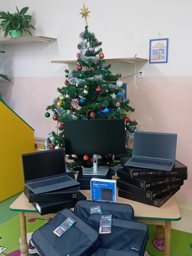 Choinka stojąca w centrum pokoju, ozdobiona bombkami, łańcuchami i gwiazdą na czubku. Przed choinką na niskim stole leżą monitor, dwa otwarte laptopy i kilka zapakowanych pudełek. Na pierwszym planie trzy czarne torby na laptopy z metkami. Wnętrze z jasnoróżową ścianą, półkami i doniczką na górze.