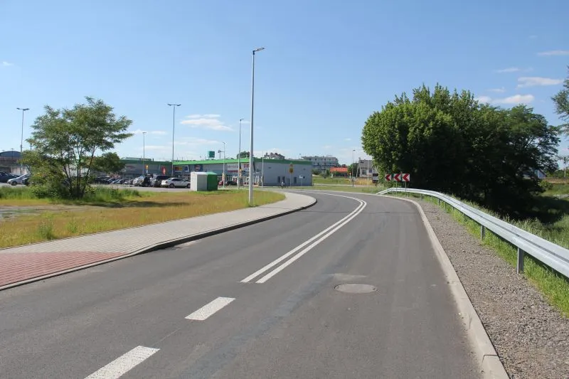 Dwupasmowa droga z chodnikiem po lewej stronie, latarniami i znakami ostrzegawczymi na zakręcie.