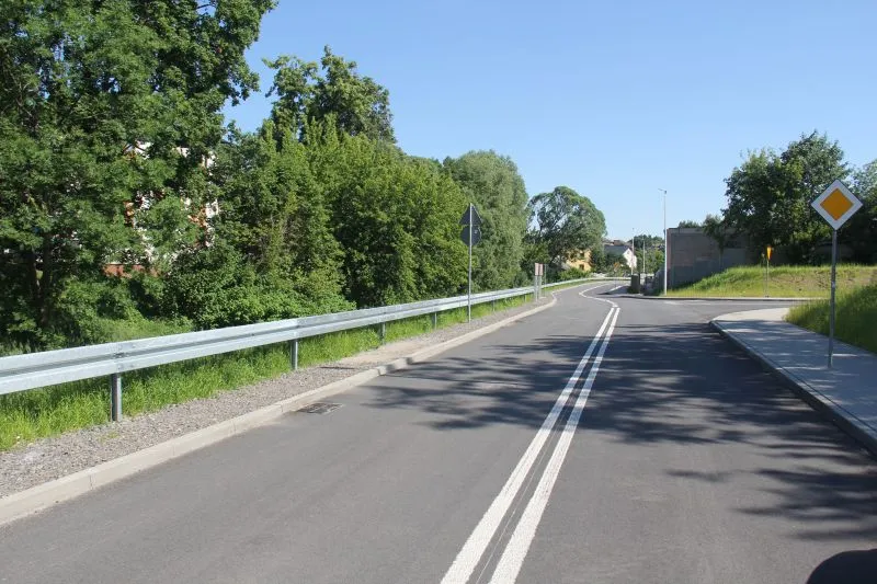 Pusta dwupasmowa droga z podwójną linią ciągłą, metalową barierką i znakami drogowymi po obu stronach, otoczona zielonymi drzewami.