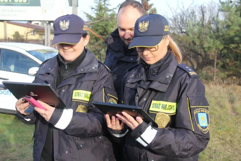 Dwóch funkcjonariuszy Straży Miejskiej w mundurach korzysta z tabletów, obok stoi mężczyzna w czarnej kurtce.