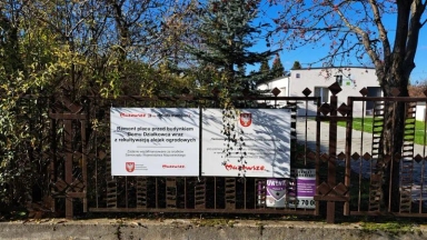 Metalowy płot z zamocowanymi dwoma tablicami informacyjnymi i budynkiem w tle.