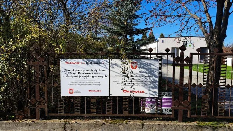 Metalowy płot z zamocowanymi dwoma tablicami informacyjnymi i budynkiem w tle.