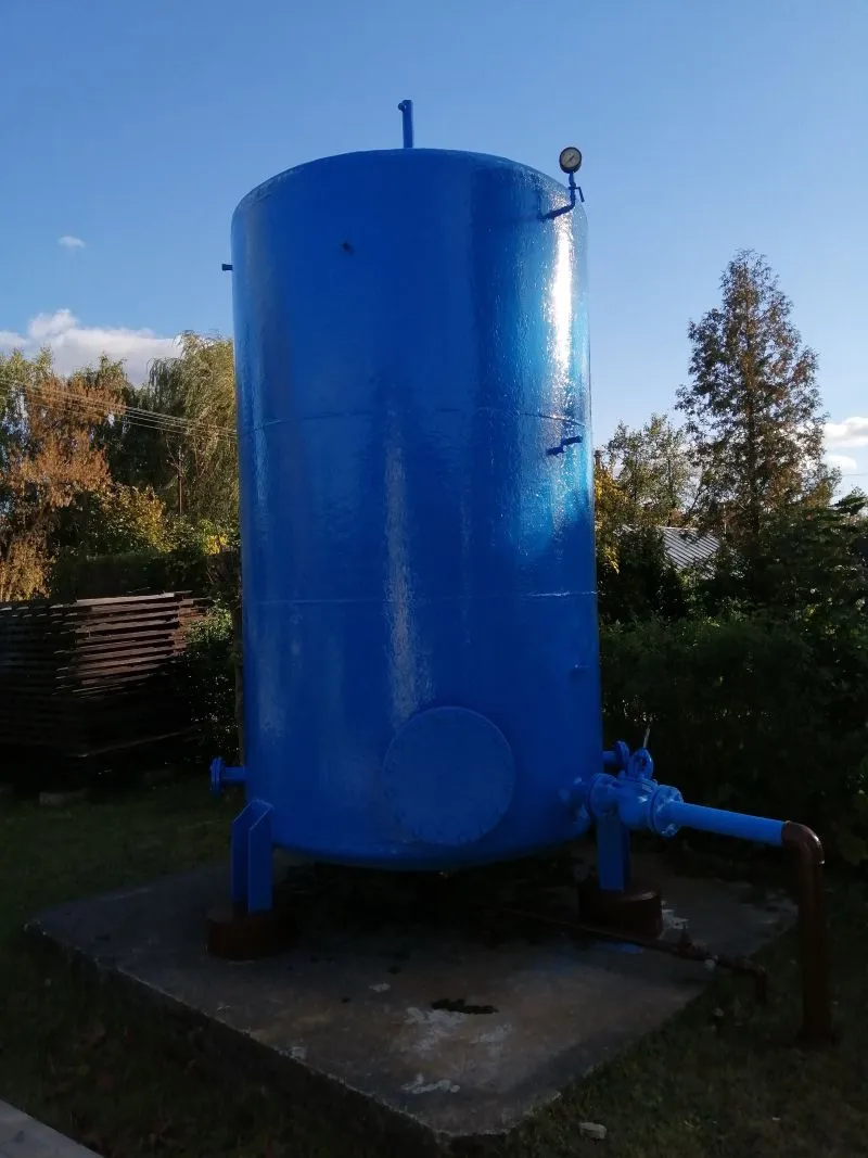 Niebieski zbiornik ciśnieniowy z manometrem i rurą na betonowej podstawie na zewnątrz.