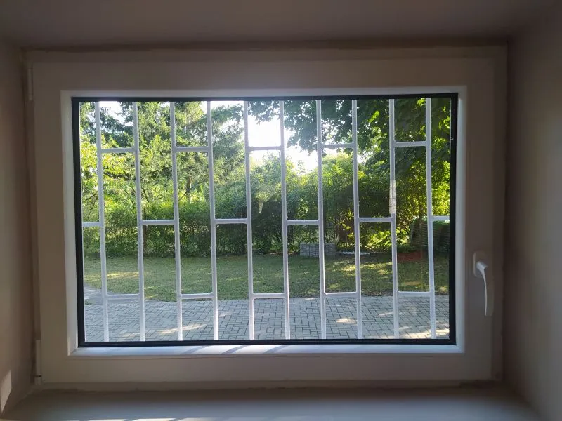 Okno z metalową kratą, przez które widać chodnik i zadrzewiony trawnik.