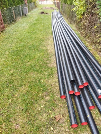 Długi stos czarnych rur z czerwonymi końcówkami leżących na trawie między dwoma ogrodzeniami.