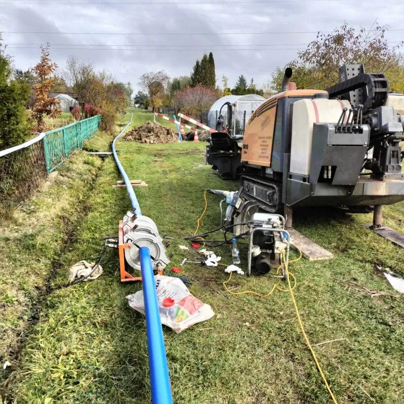 Maszyna budowlana i niebieska rura ułożona na trawie obok ogrodzenia i wykopu.