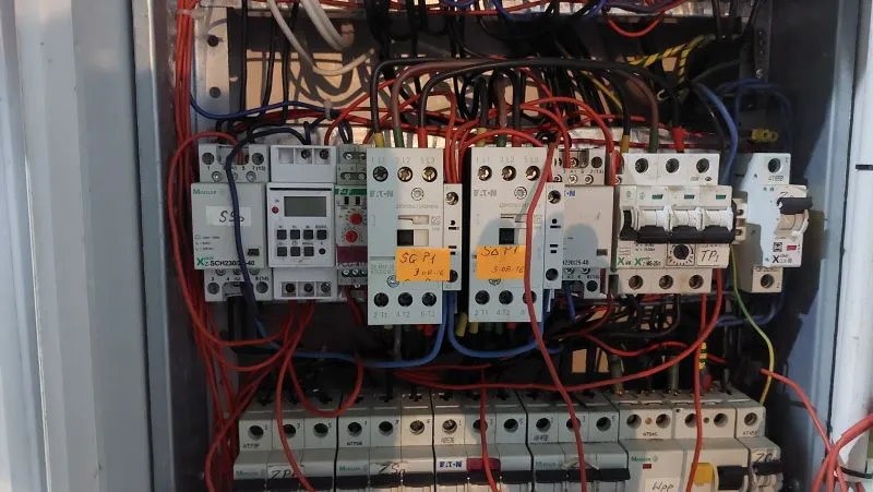 Moduły elektryczne, przekaźniki i wyłączniki z licznymi czerwonymi i niebieskimi przewodami podłączonymi w szafce elektrycznej.