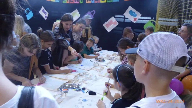 Grupa dzieci i dorosłych siedzi wokół stołu podczas zajęć plastycznych.