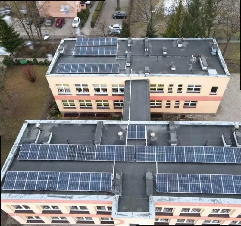 Dwa budynki połączone łącznikiem z zamontowanymi panelami fotowoltaicznymi na płaskich dachach.