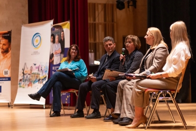 Pięć osób siedzi na krzesłach na scenie podczas panelu dyskusyjnego, jedna z kobiet mówi do mikrofonu.