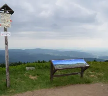 Tablica informacyjna i drewniany słup z drogowskazami stoją na górskim szlaku z widokiem na rozległe pasmo górskie.