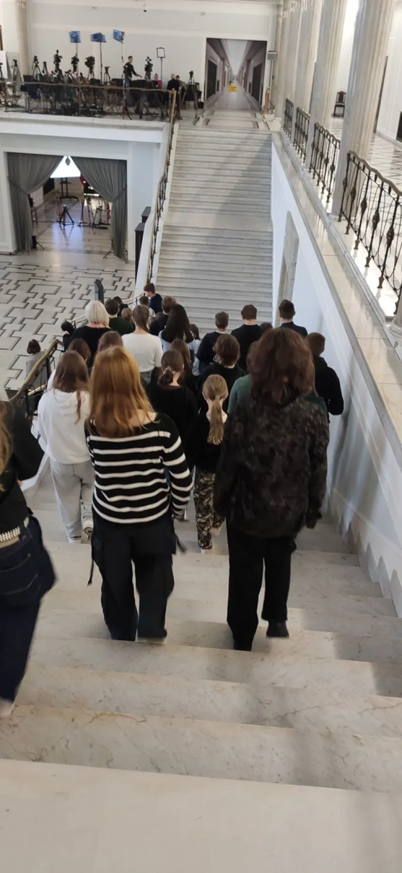 Grupa osób schodzi po marmurowych schodach w przestronnym wnętrzu z białymi kolumnami i balustradami.