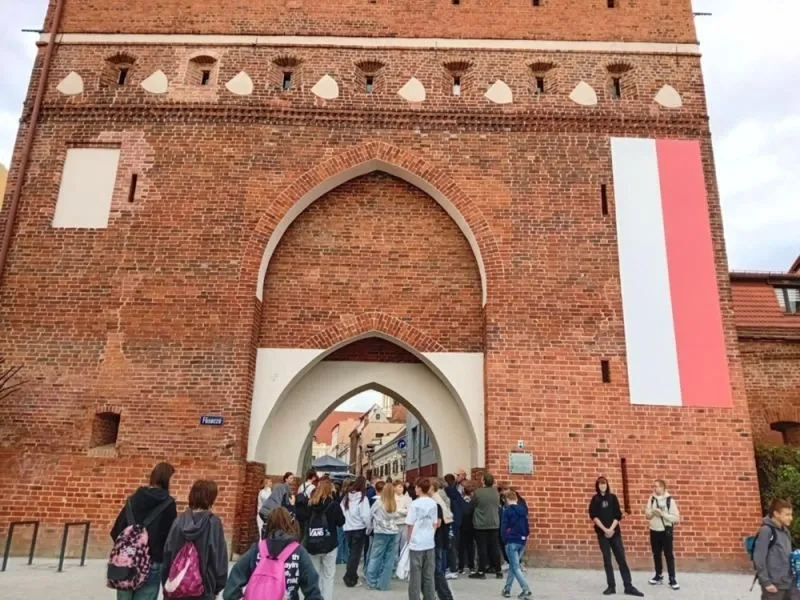 Grupa ludzi stoi przy czerwonej ceglanej bramie z dwoma ostrołukowymi wejściami oraz pionowym pasem biało-czerwonym po prawej stronie.